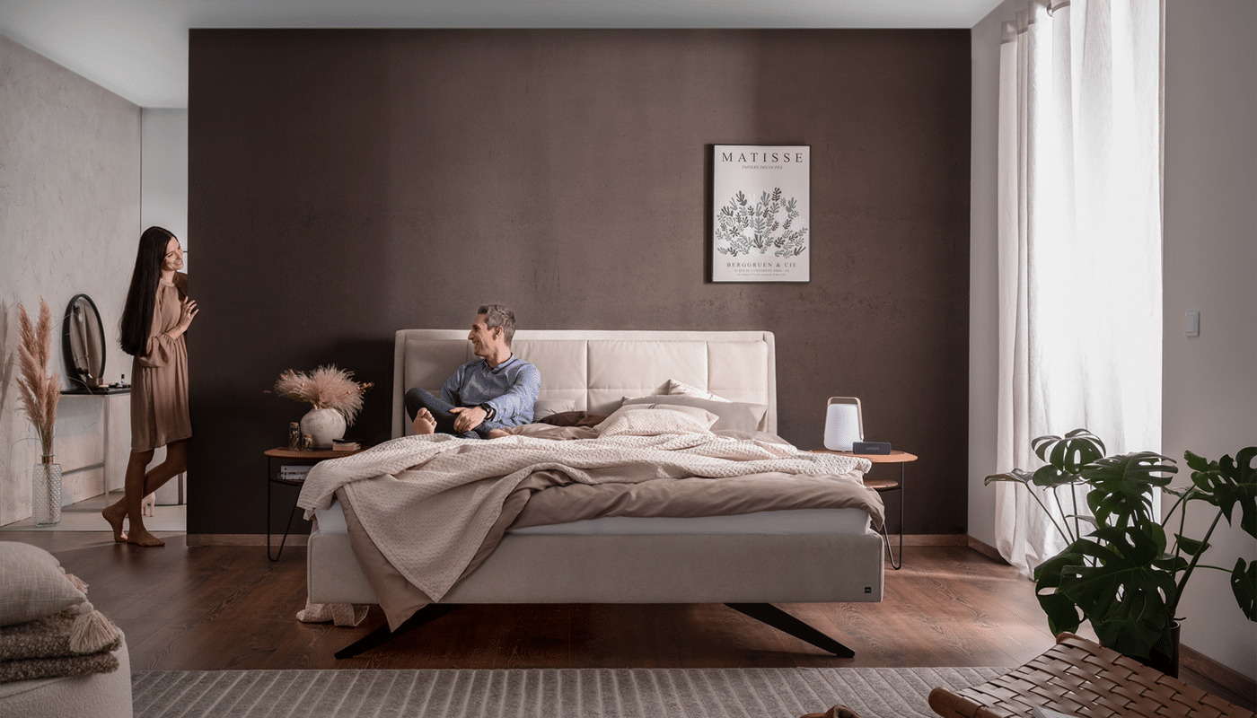 Bett Decuro in hellgrauem Stoff mit Wendekissen im gemütlich gestaltetet Schlafzimmer mit einem Paar, das sich unterhält Bett Decuro in hellgrauem Stoff mit Wendekissen im gemütlich gestaltetet Schlafzimmer mit einem Paar, das sich unterhält