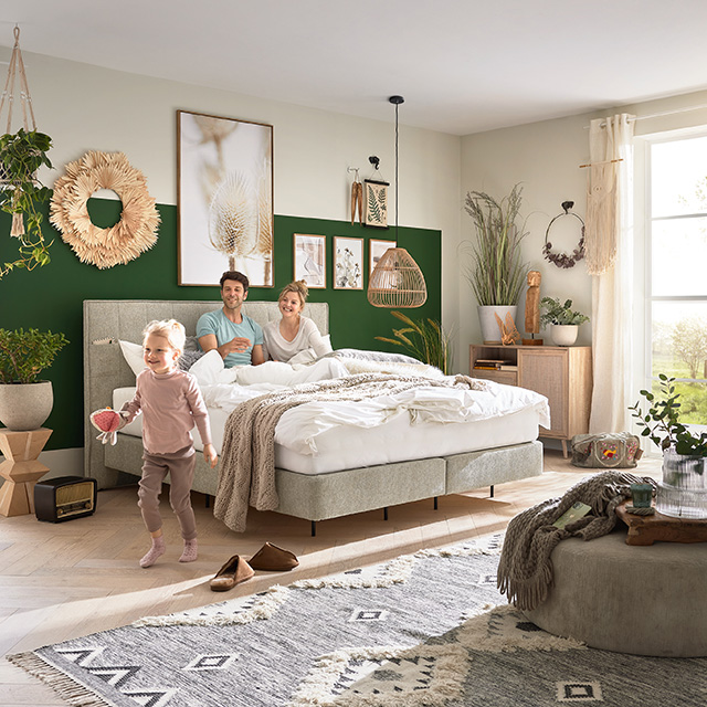 Paar sitzt im Bett, Kind kommt dazu in einem modernen Schlafzimmer im Boho-Stil