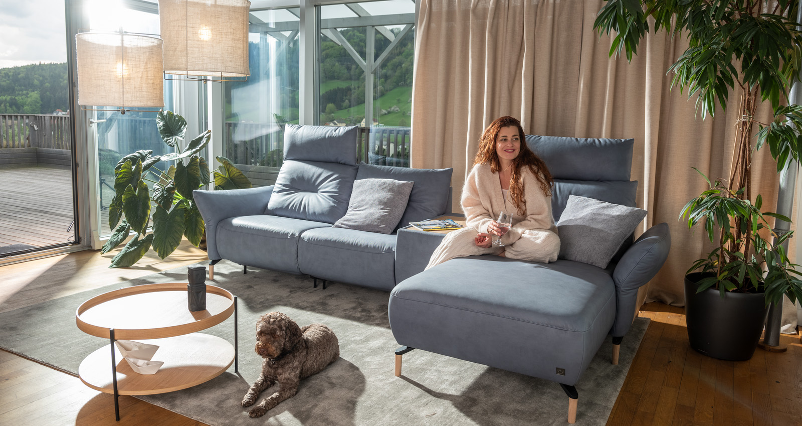 Frau sitzt mit Wasserglas auf dem Ecksofa, ein Hund liegt davor auf dem Teppich