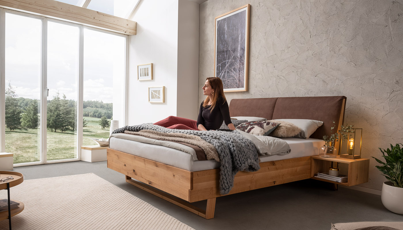 Frau sitzt entspannt auf einem modernen Massivholzbett in einem hellen Schlafzimmer mit Ausblick ins Grüne.