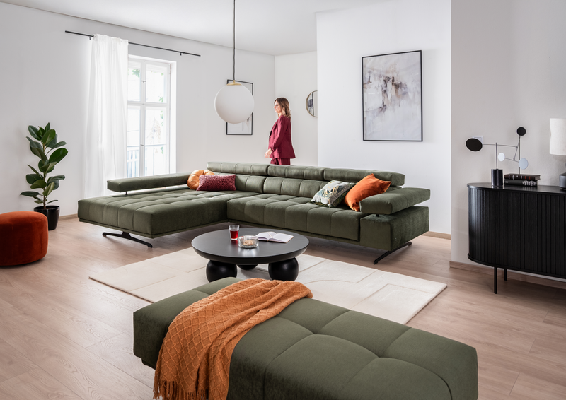 xl-Raimo Eine Frau geht hindert dem Sofa RAIMO durch das Wohnzimmer